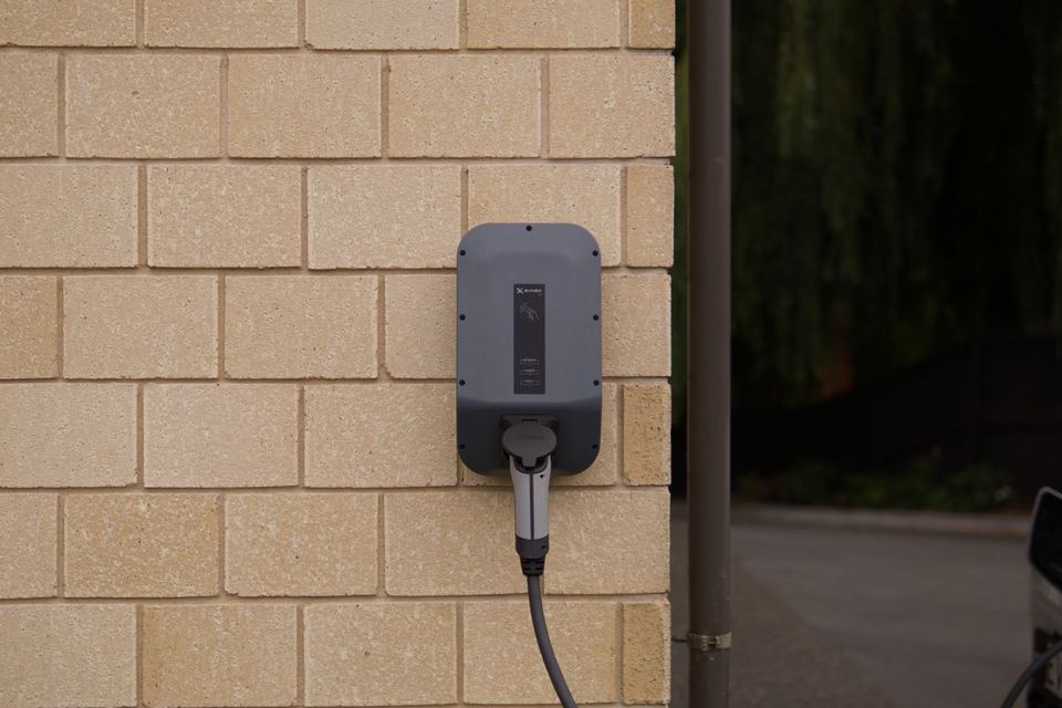 Carregador veicular (wallbox) fixado em parede de tijolos, com cabo conectado — imagem ilustrativa de banco (Ed Harvey / Pexels), não é instalação da E5